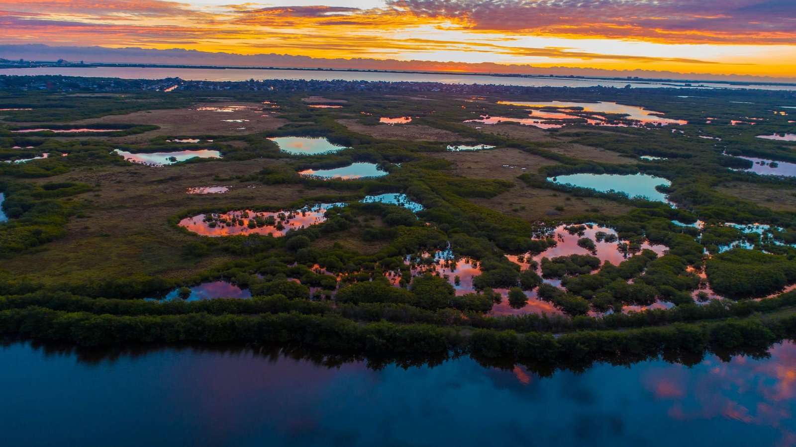 Aerial view of a coastal wetland at sunset, scattered ponds reflecting an orange and pink sky, dark open water in the foreground.